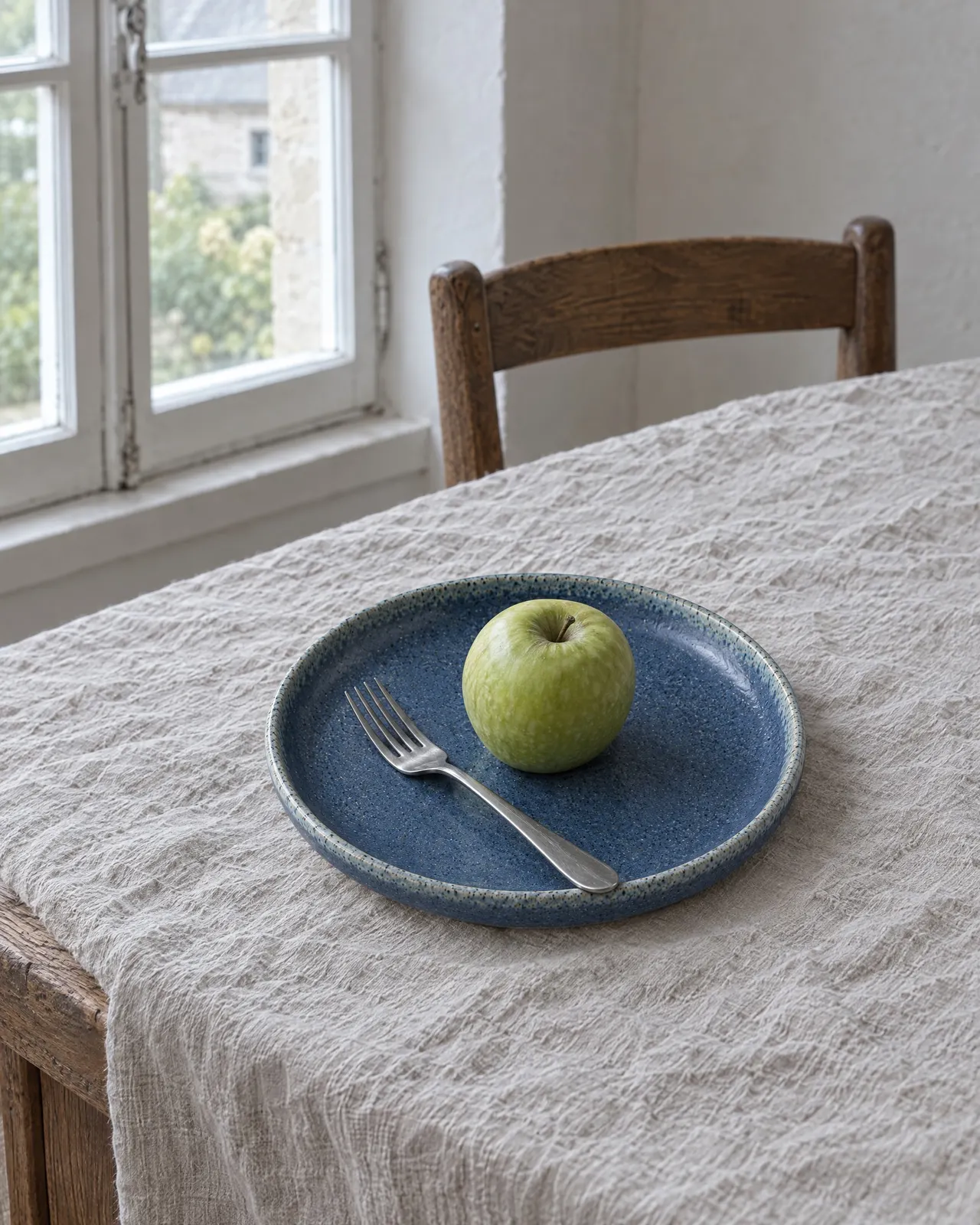 Assiette en grès bleu posée sur une nappe en lin, une pomme verte et une fourchette dessus