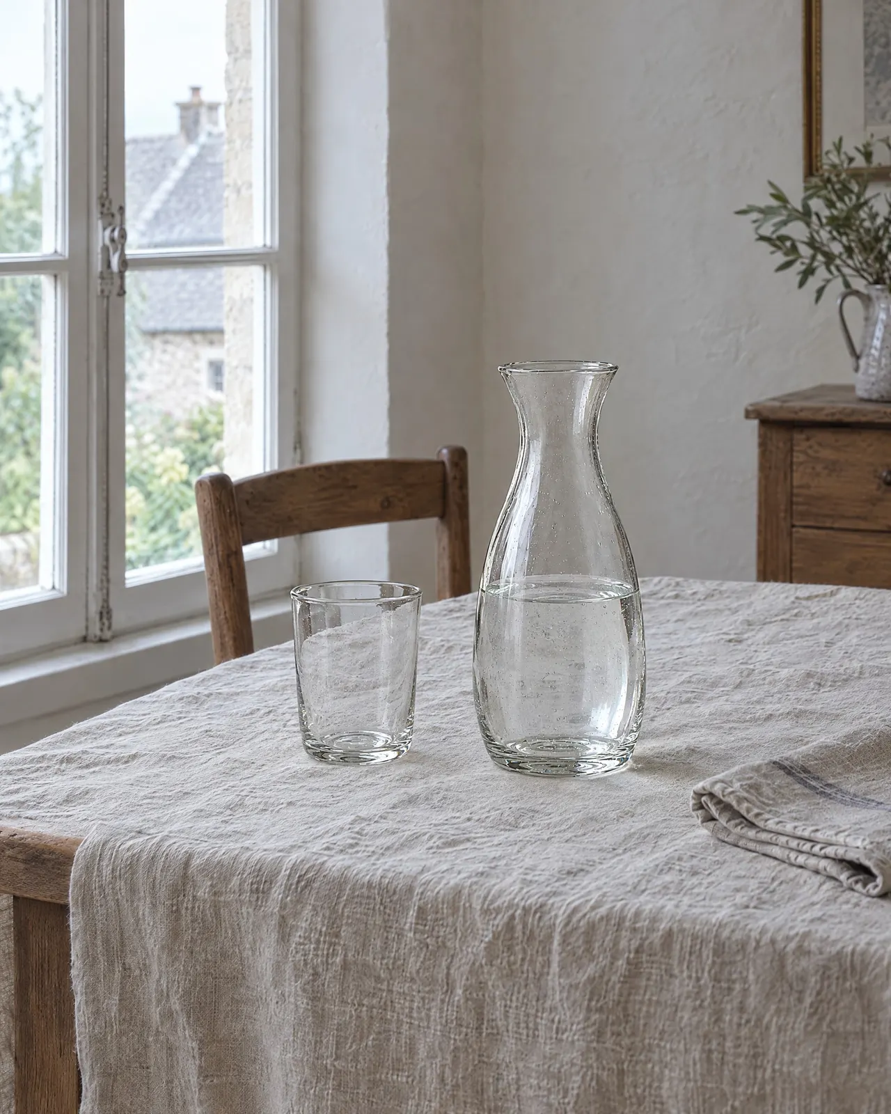 Carafe en verre soufflé remplie d'eau, posée sur une nappe en lin à côté d'un verre, près de la fenêtre