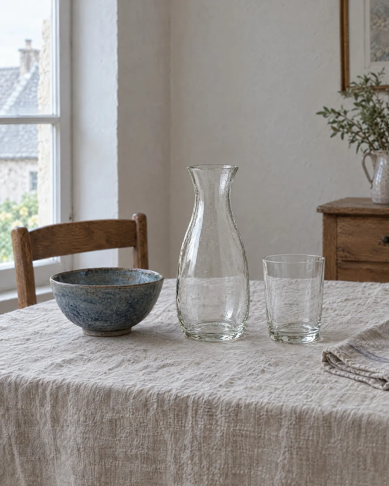 Carafe en verre soufflé posée sur une nappe en lin, entre un bol en grès bleu et un verre