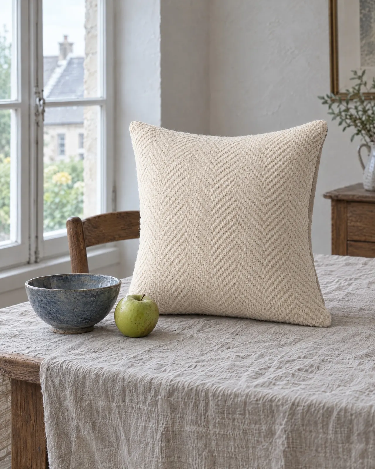 Coussin en laine tissée écrue posé sur une nappe en lin, près d'un bol bleu et d'une pomme verte