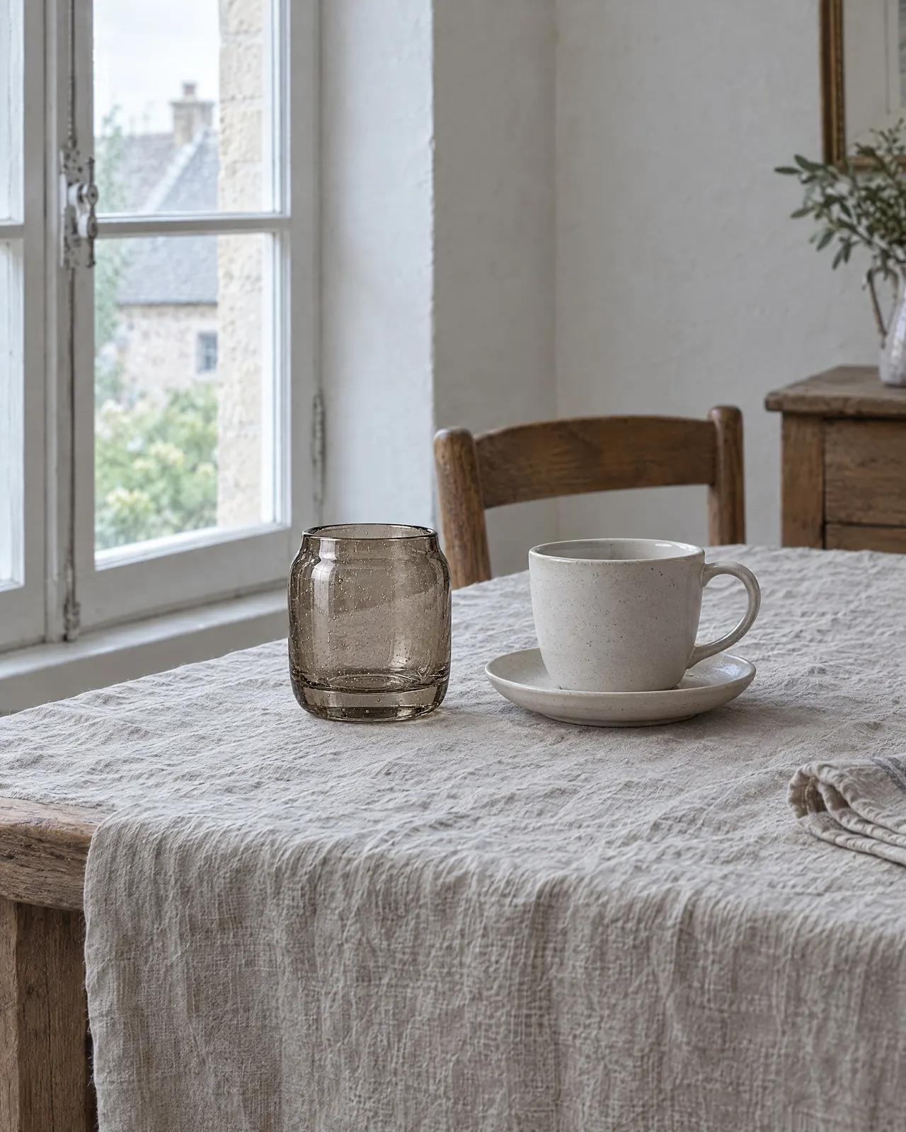 Photophore en verre fumé posé sur une nappe en lin, près d'une tasse en grès sable sur sa soucoupe