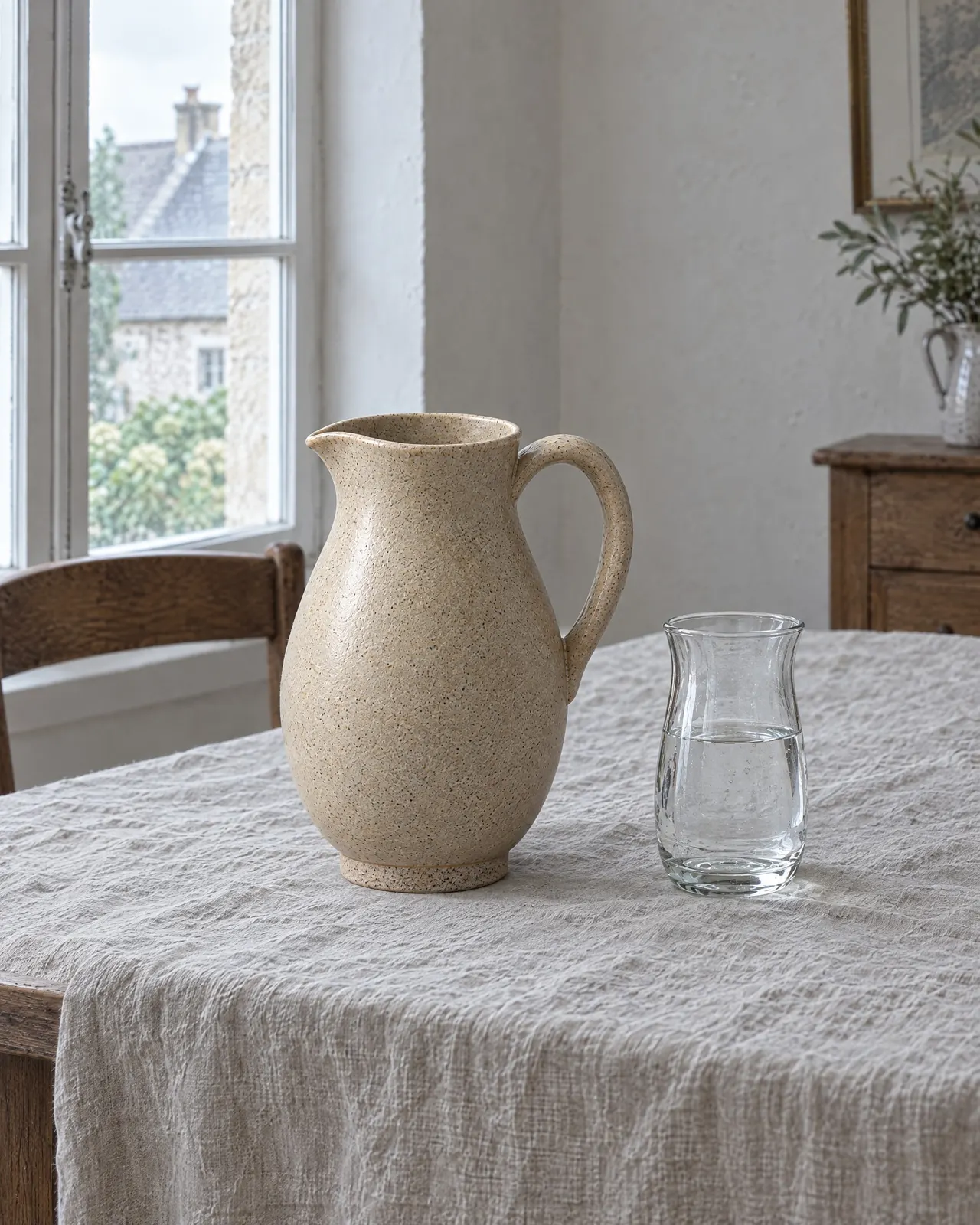 Pichet en grès sable posé sur une nappe en lin à côté d'un verre à eau, près de la fenêtre