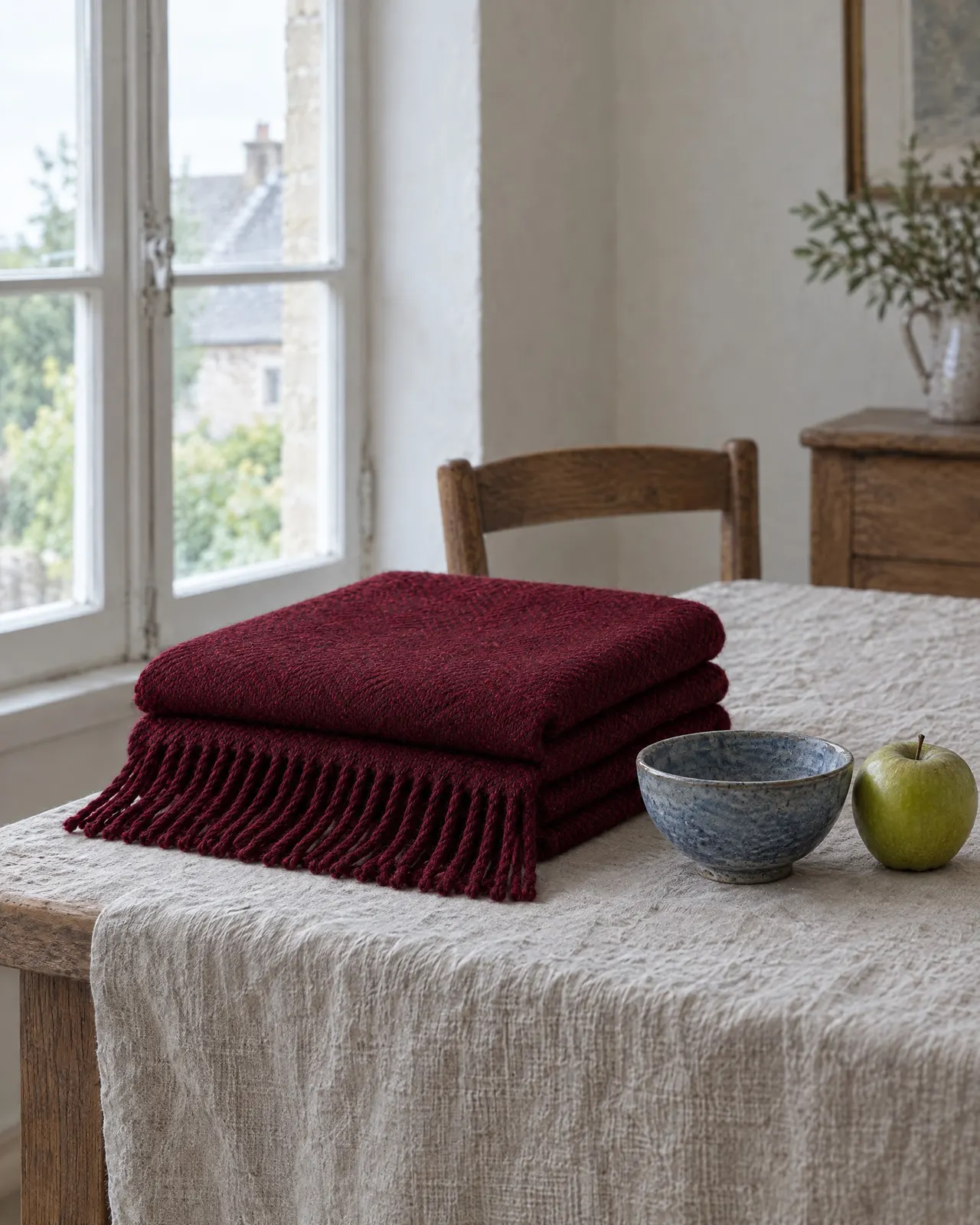 Plaid en laine grenat plié sur une nappe en lin, près d'un bol bleu et d'une pomme verte