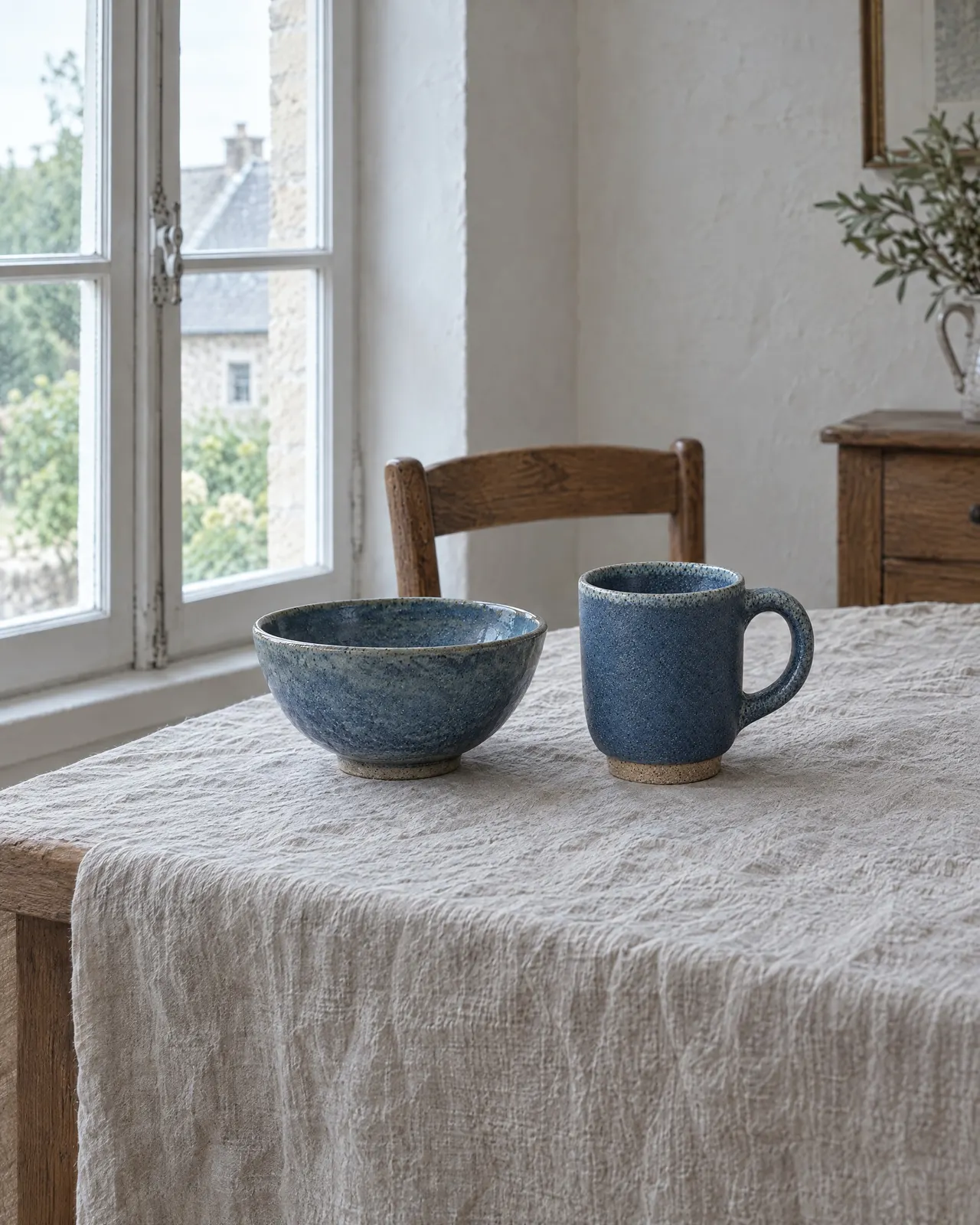 Tasse en grès bleu posée à côté du bol assorti sur une nappe en lin, près de la fenêtre