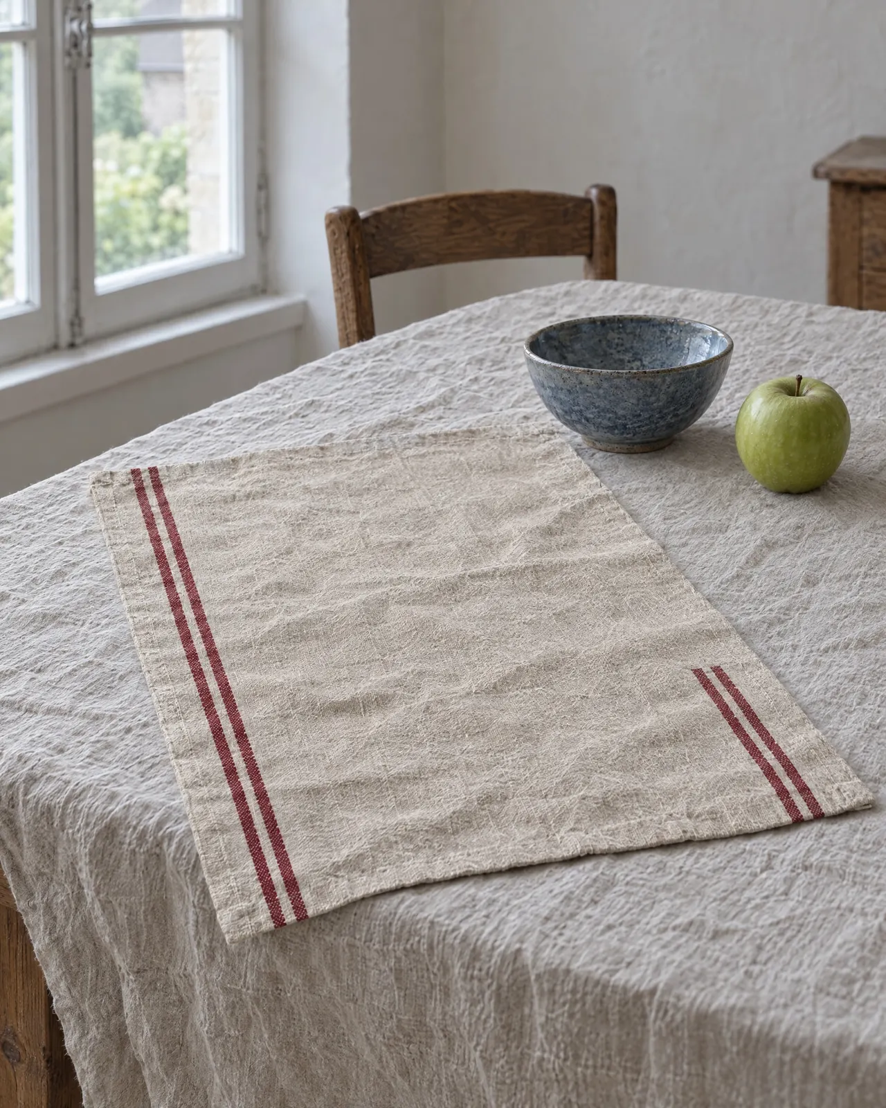Torchon en lin rayé rouge déplié sur une nappe en lin, près d'un bol bleu et d'une pomme verte